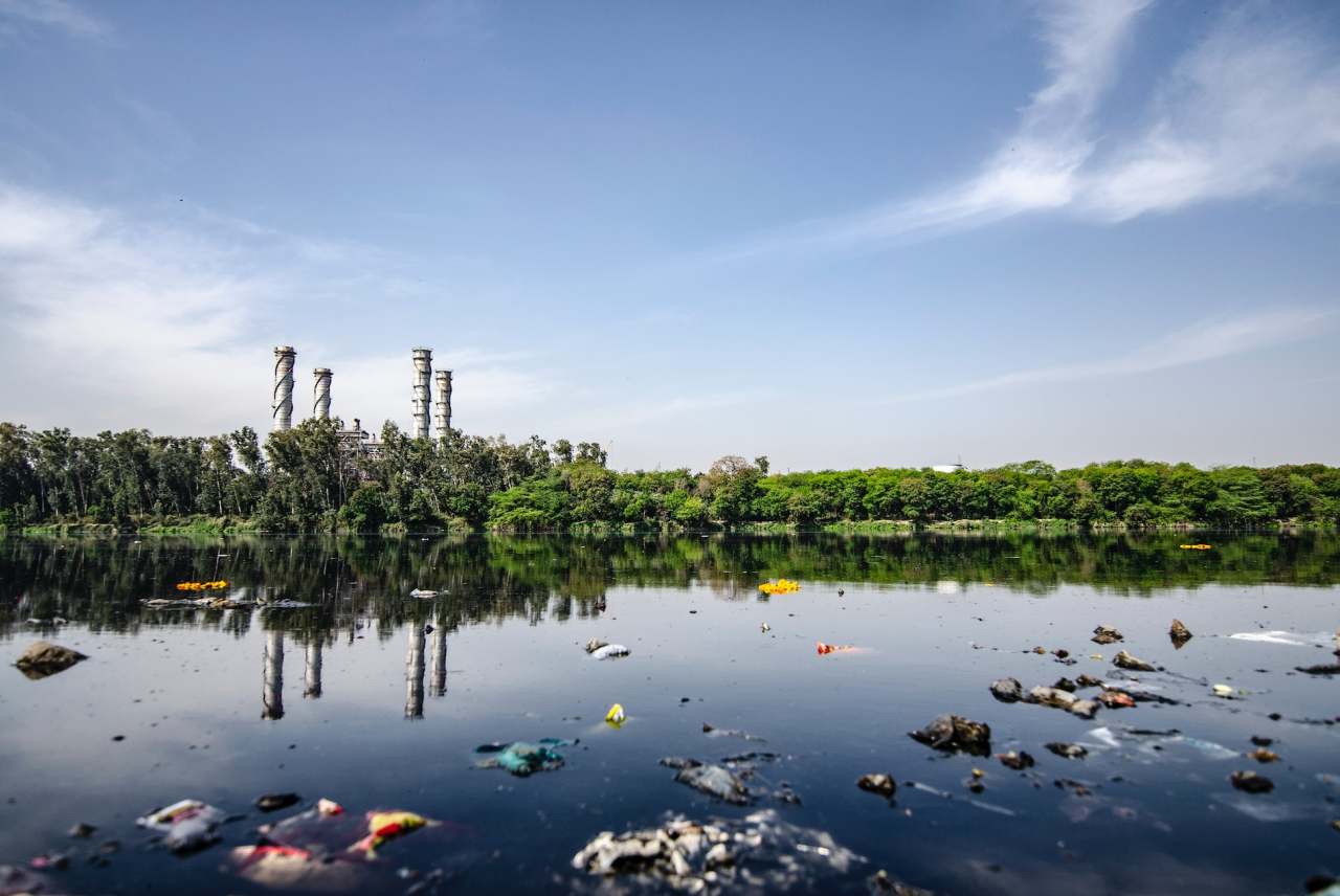 Industrial plant among trees with rubbish in water in foreground